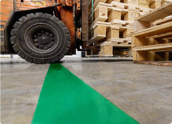 En stor gaffeltruck kjører over en linje med grønn ToughStripe Max gulvmerkingstape. Kantene på tapen er uskadd til tross for gaffeltruckens tunge hjul.