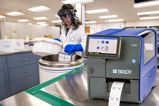 A lab technician works with in a laboratory, while a Brady i7500 label printer prints labels on a nearby counter.